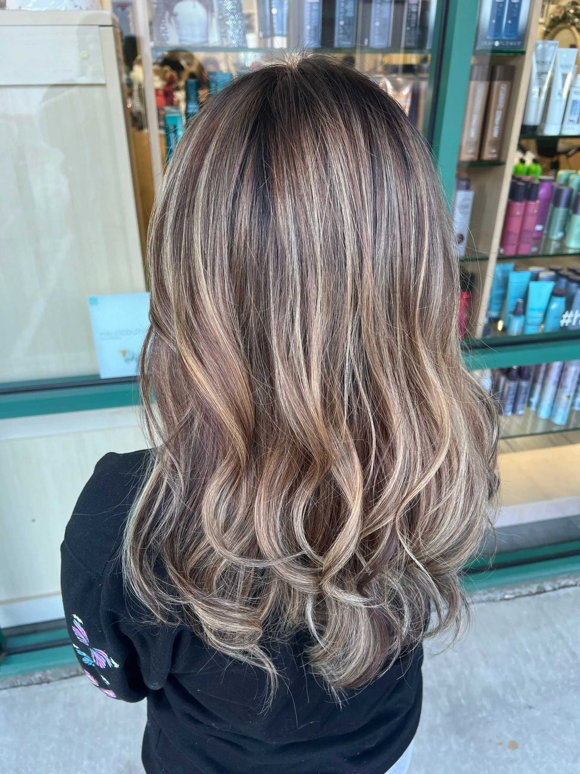 Woman's brown and blonde highlighted wavy hair. She's wearing a black top. Background shows beauty products.