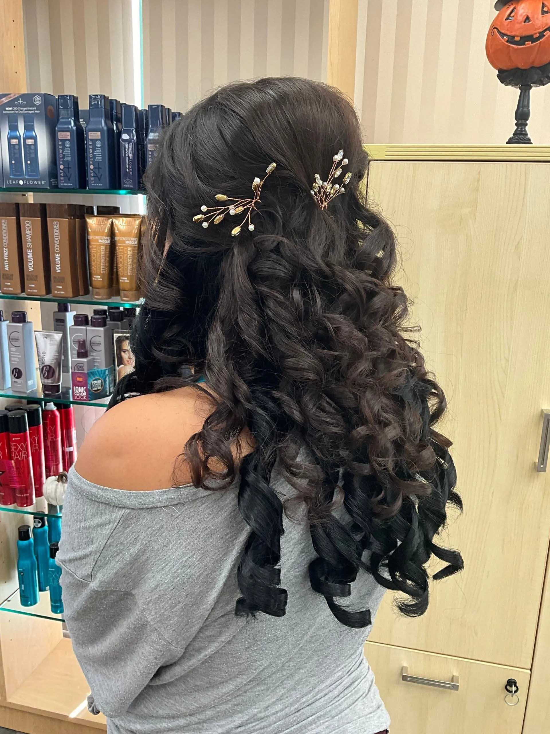 Woman with curly dark hair, half-updo style, gold hair accessories, gray top. Salon setting.