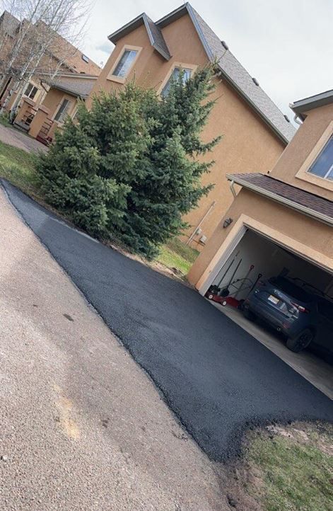 Black asphalt driveway leads up to a tan stucco house with an open garage; a car is parked inside.