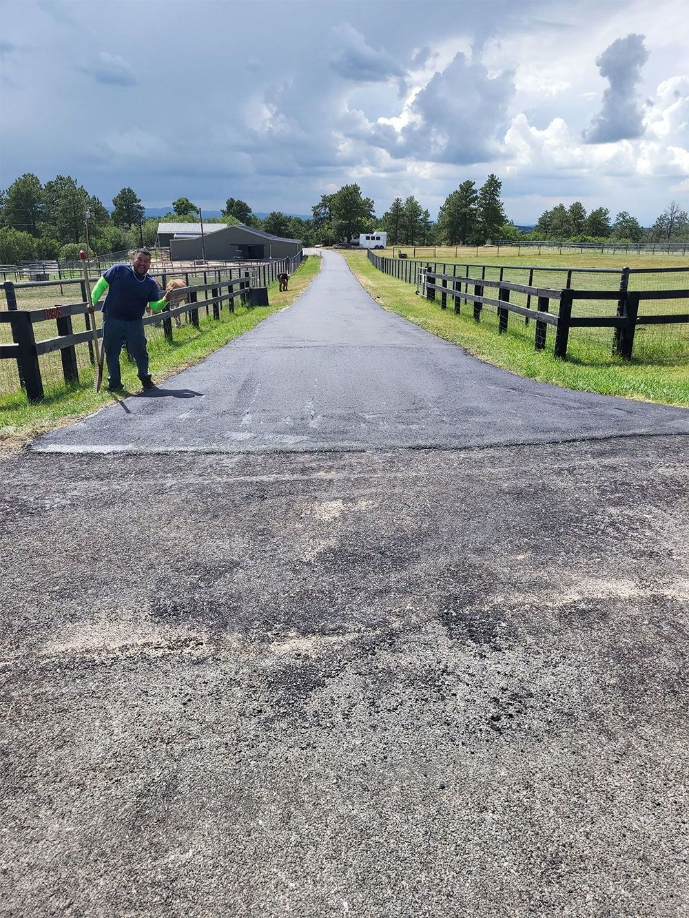 Person spreading gravel on a long, paved driveway lined with fences, under a cloudy sky.