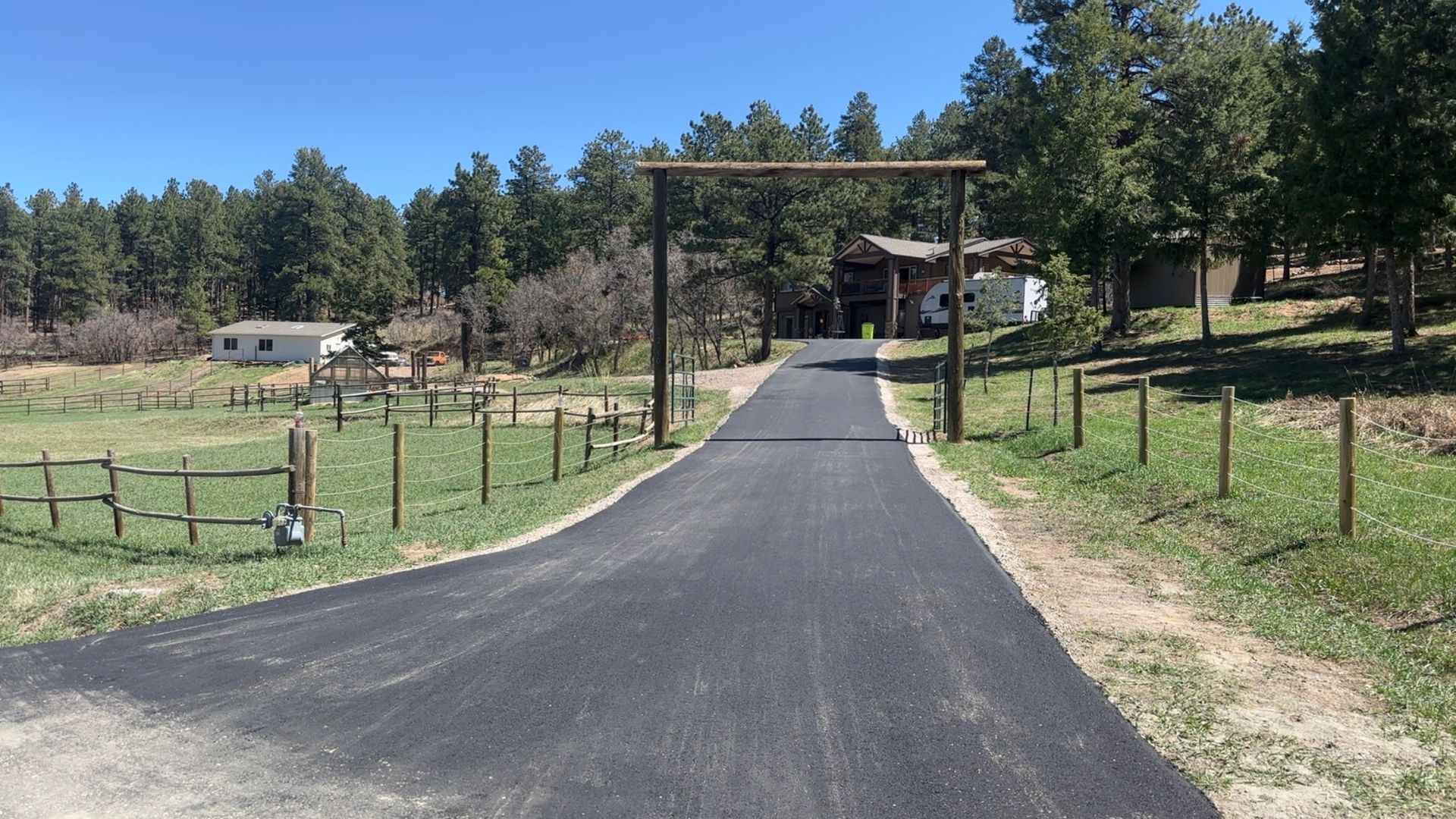 Paved driveway leads to a wooden archway and house, flanked by fields and trees under a blue sky.