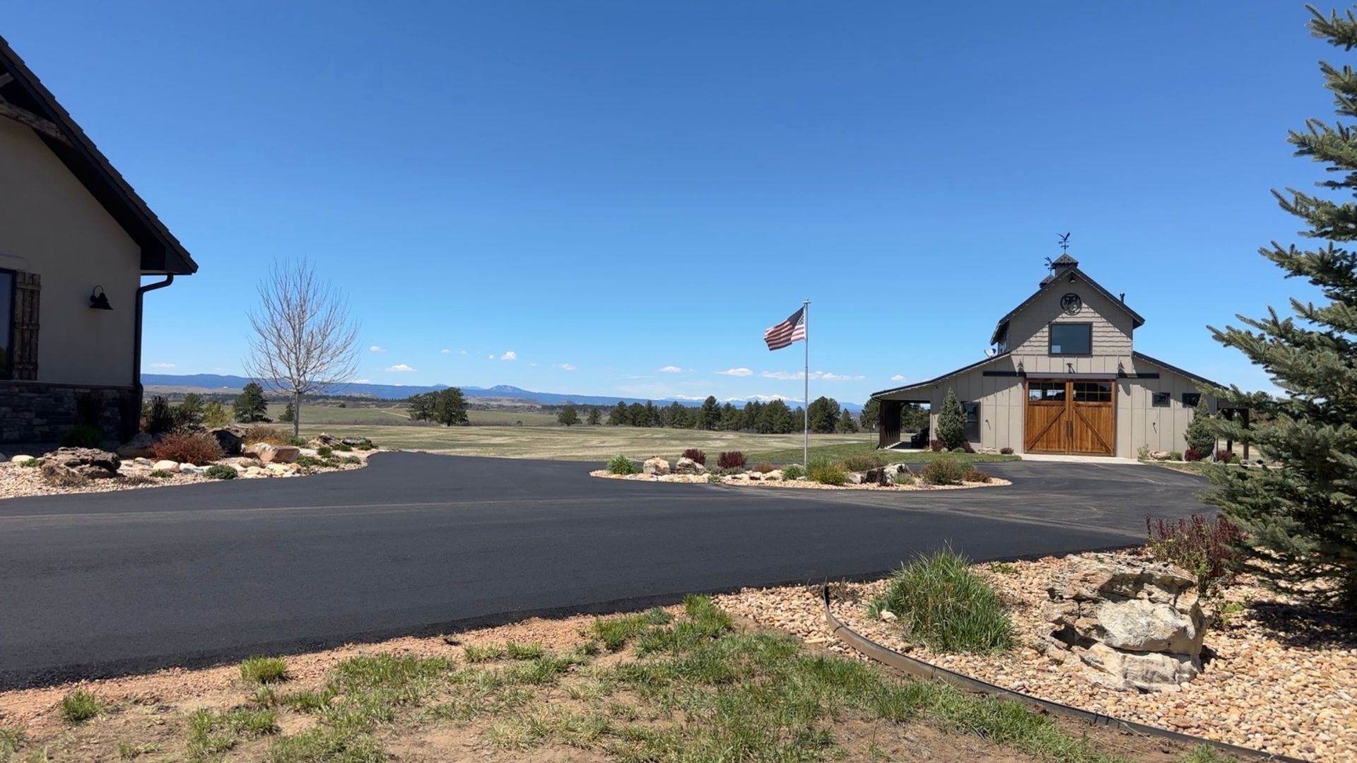 A driveway leads to a barn and house under a blue sky, with a flag and distant mountains.