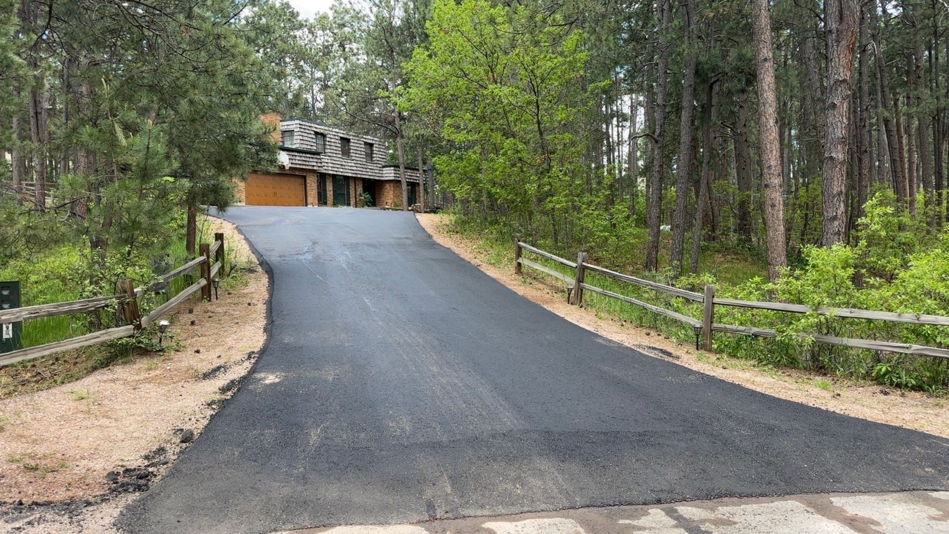 Paved driveway leading uphill to a house with a wooden fence on either side, surrounded by trees.