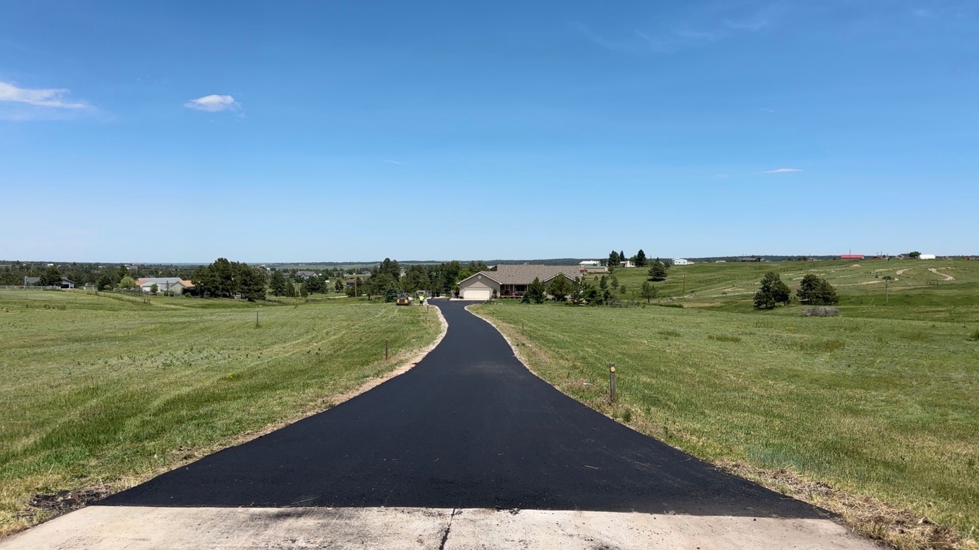 Long asphalt driveway leads to a house set in a grassy field under a blue sky.