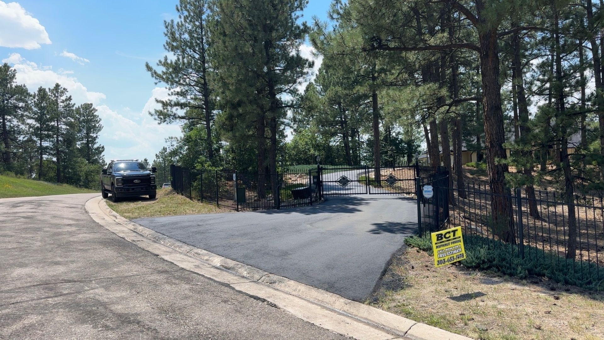 Entrance to a gated community with a black SUV parked on the left; asphalt road, trees, and blue sky.