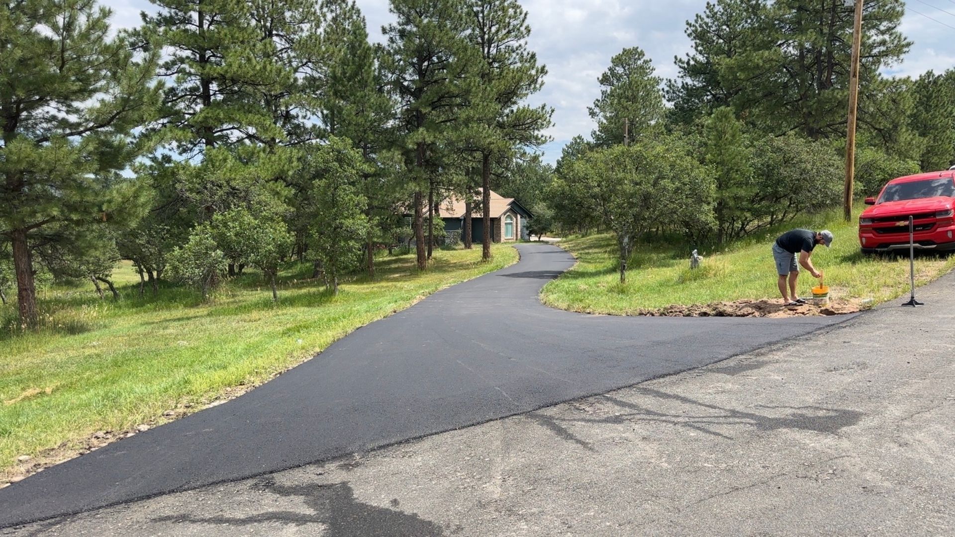 Newly paved asphalt driveway with person working near a red truck. Trees and house in the background.