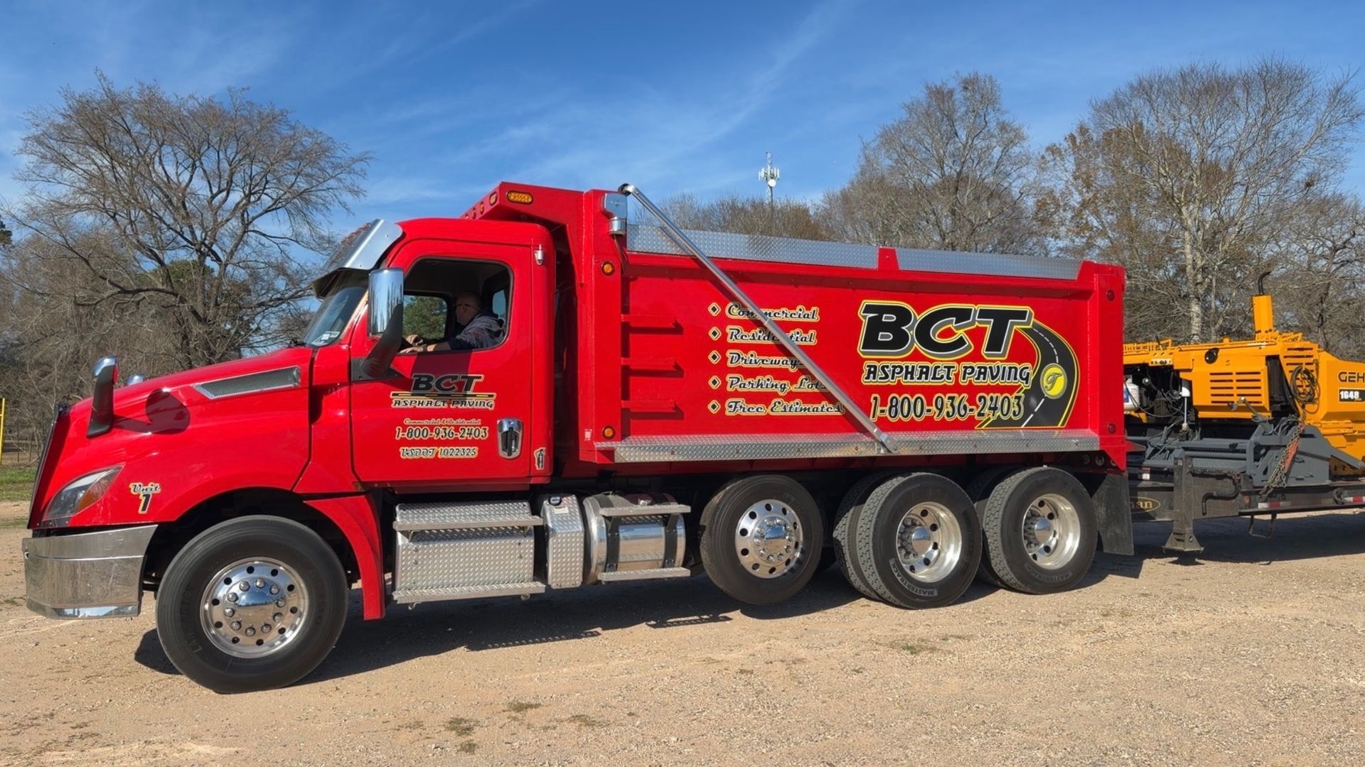 Red BCT dump truck and asphalt paver on trailer, parked outdoors.