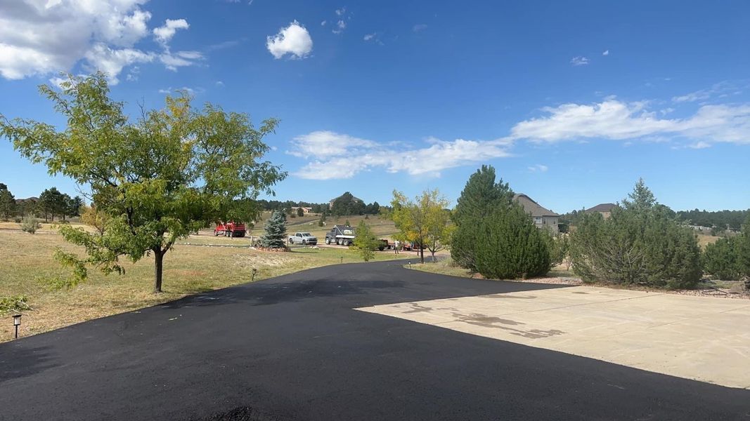 Paved driveway leads to a grassy area with trees, vehicles, and a clear blue sky with clouds.