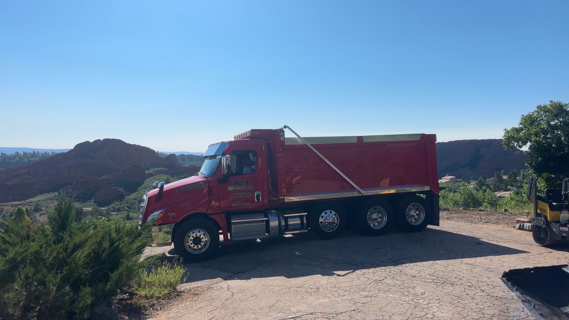 Red dump truck parked on a dirt road with a scenic rocky landscape and blue sky in the background.