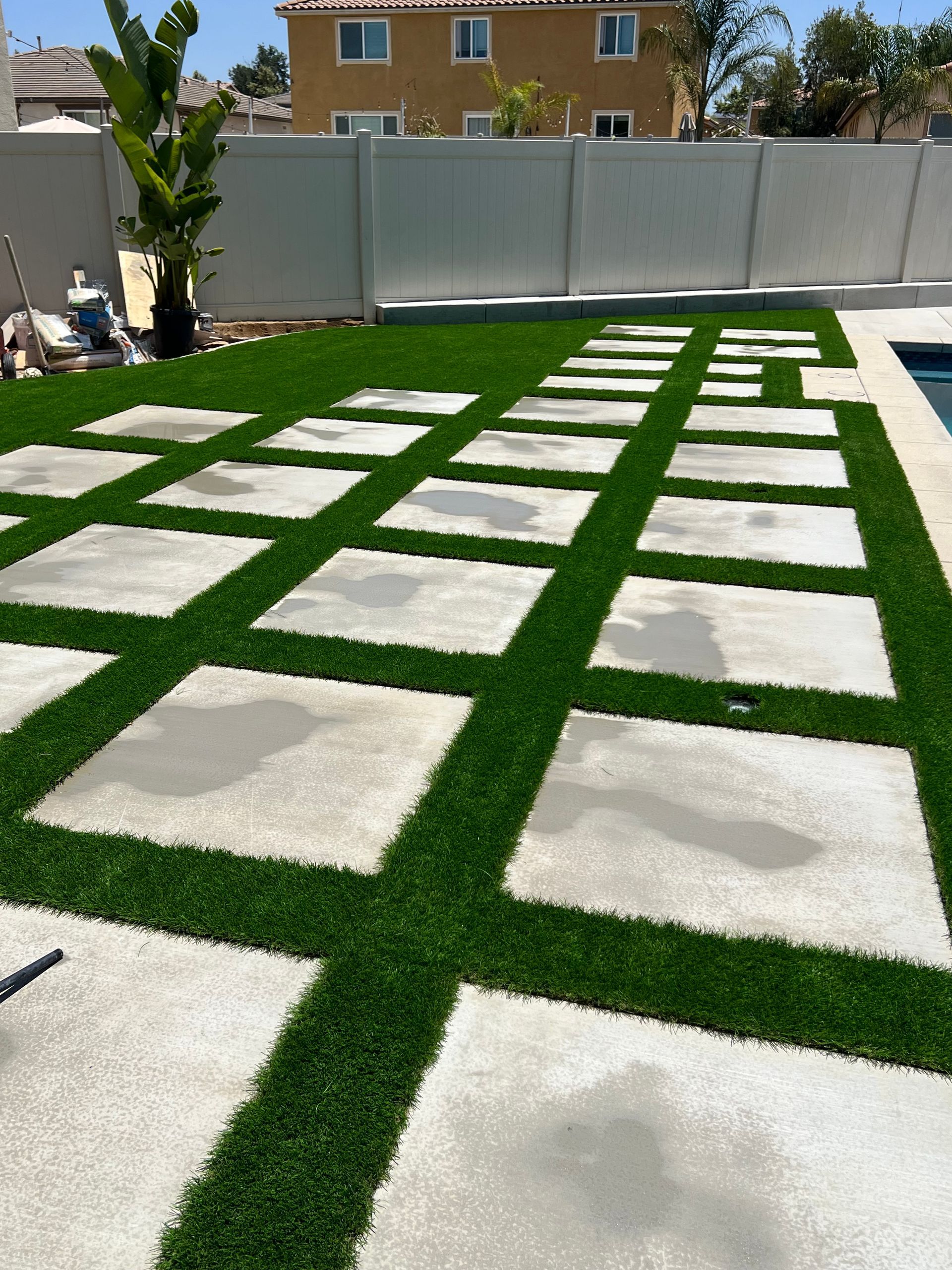 Square concrete pavers with artificial turf in between, in a backyard setting near a pool.