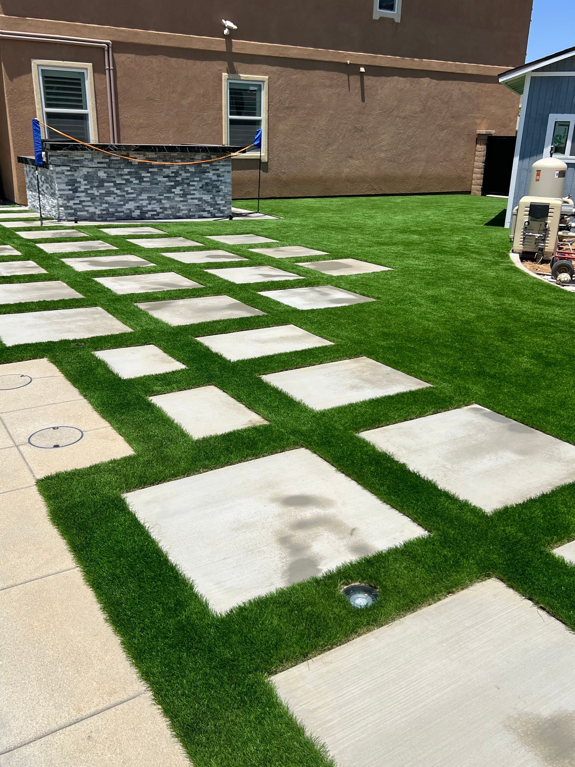 Concrete pavers set in synthetic turf, creating a walkway through a green yard. Building in background.
