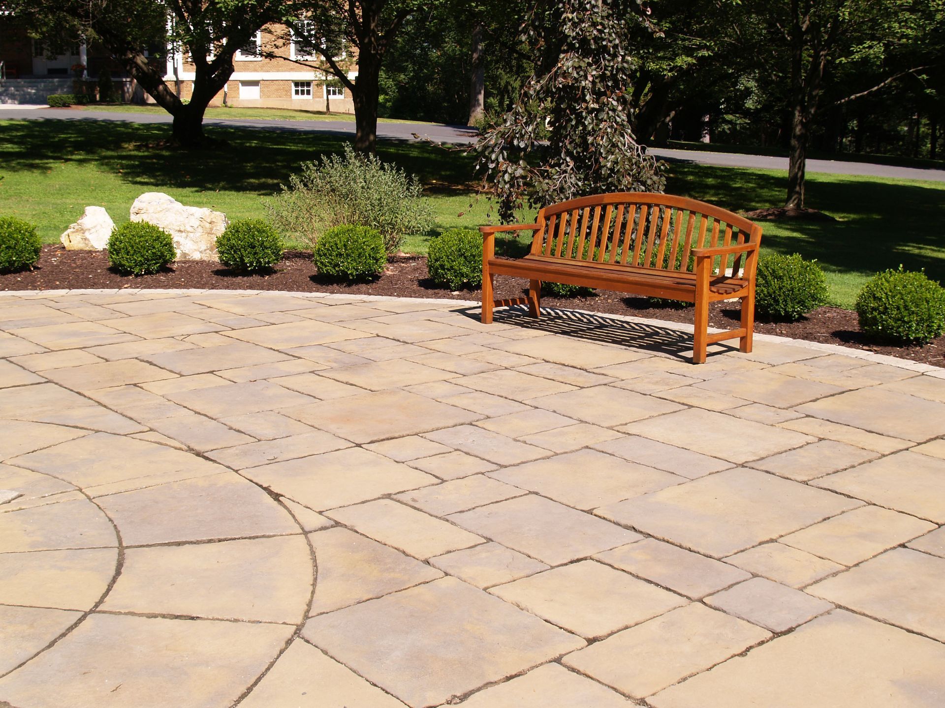 A wooden bench sits on a stone patio, surrounded by green bushes and a manicured lawn.