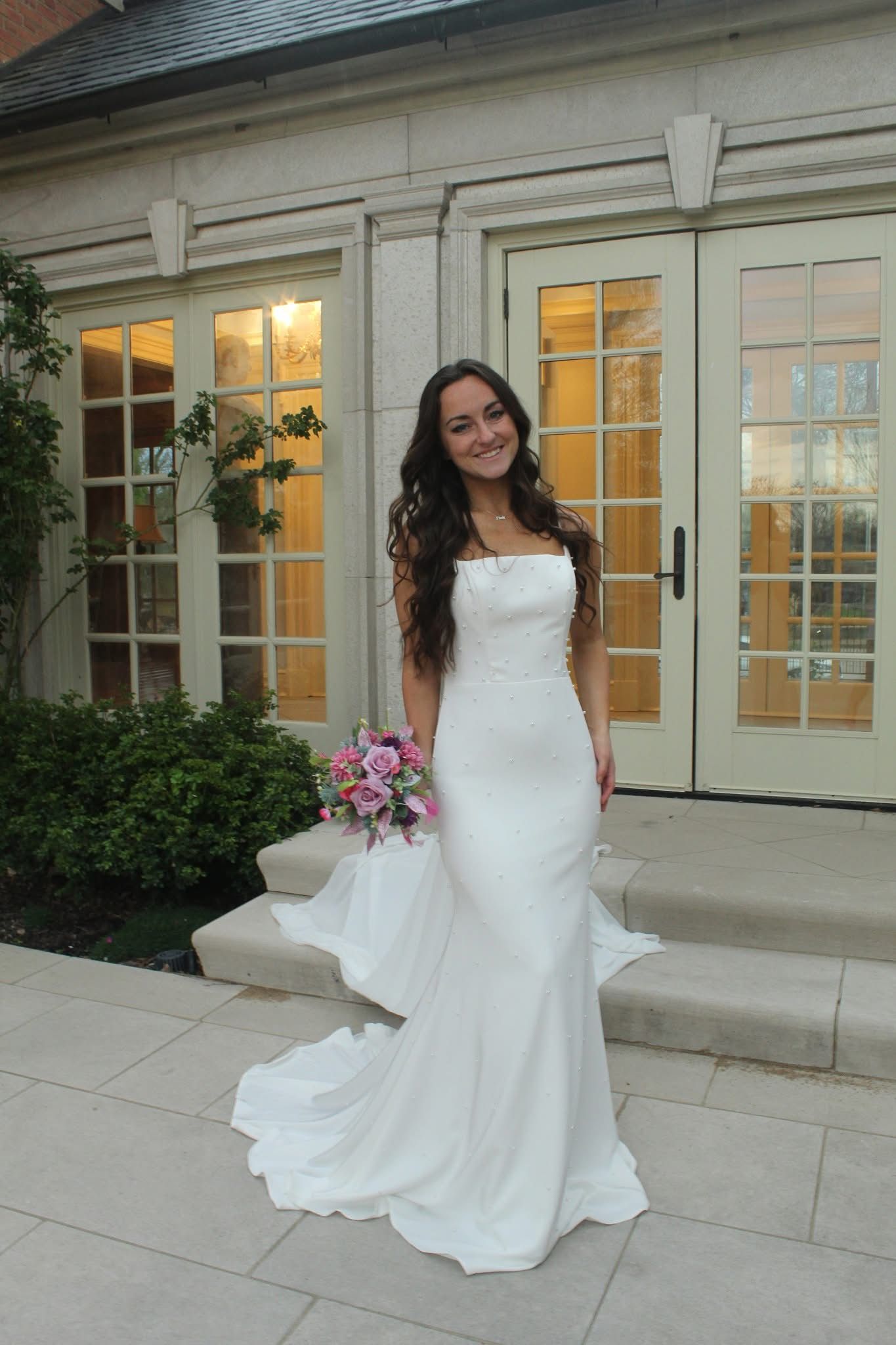 A woman in a wedding dress is standing in front of a building holding a bouquet of flowers.