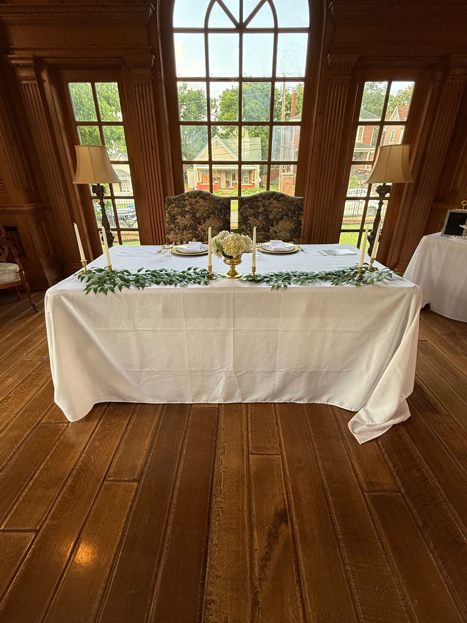 A table with a white tablecloth and candles on it in a room with a lot of windows.