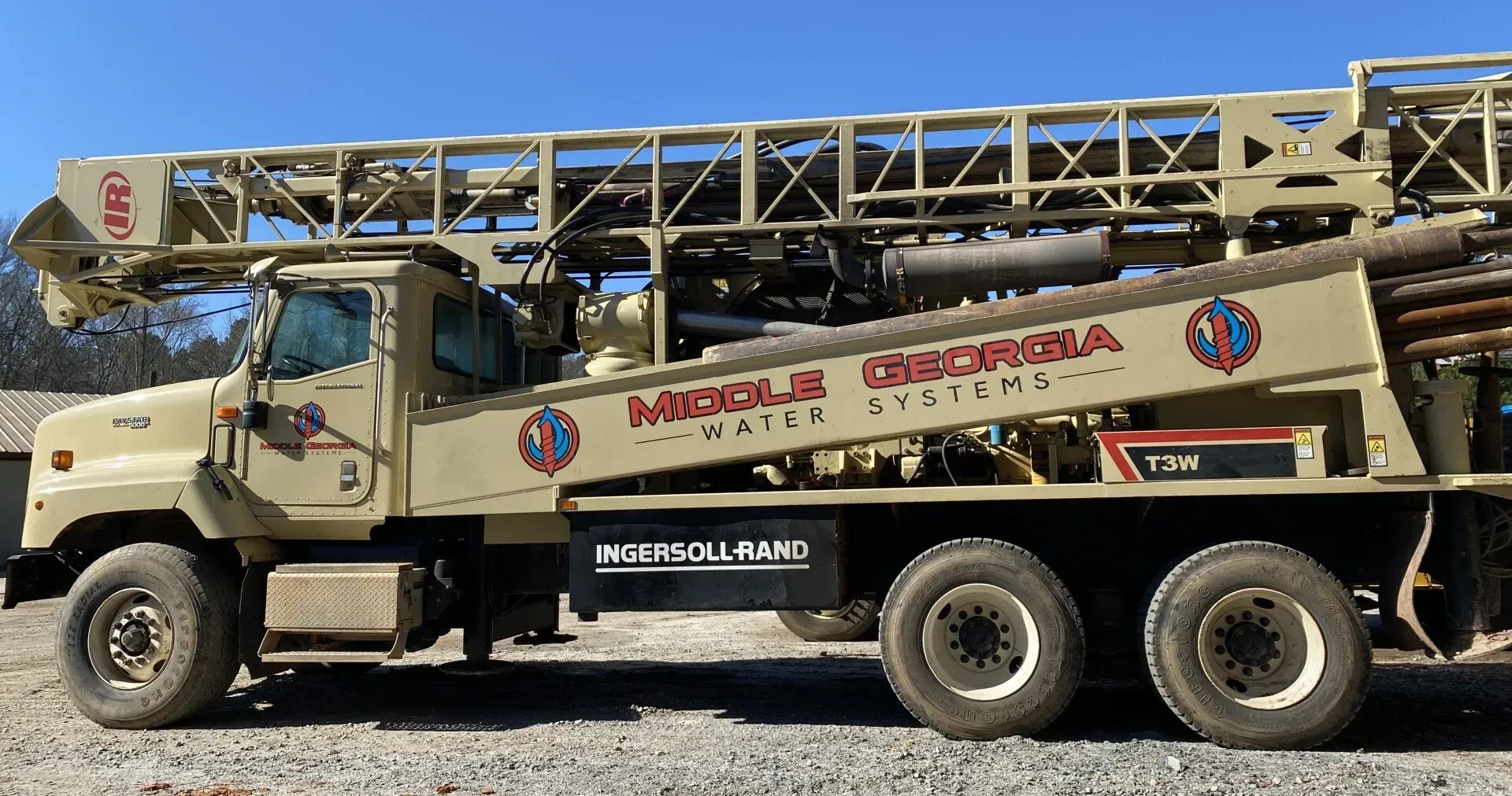 Beige utility truck with a long mounted ladder parked on gravel under a blue sky.