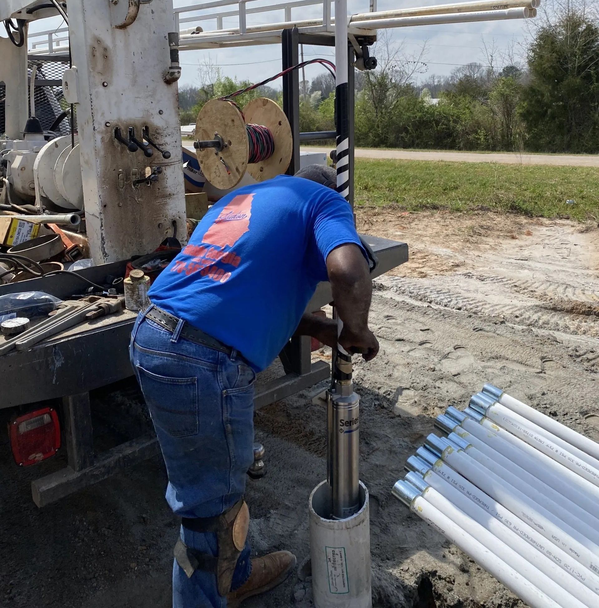 Worker in blue shirt operating drilling equipment beside stacked white PVC pipes outdoors