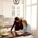 Two people looking at papers in a kitchen with white cabinets, a wood countertop, and a window.