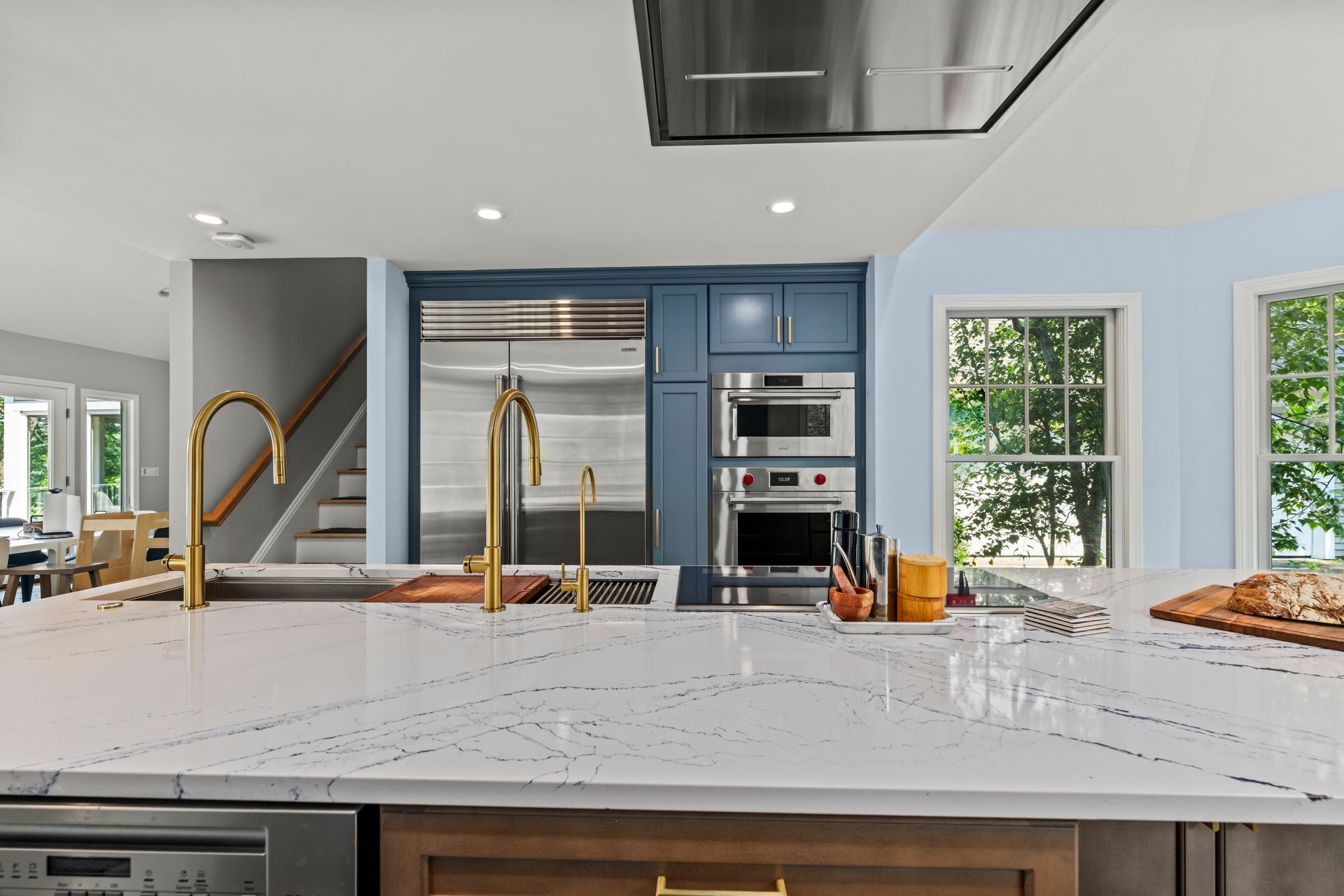 Modern kitchen with blue cabinets, gold faucets, a marble island, and large windows overlooking greenery