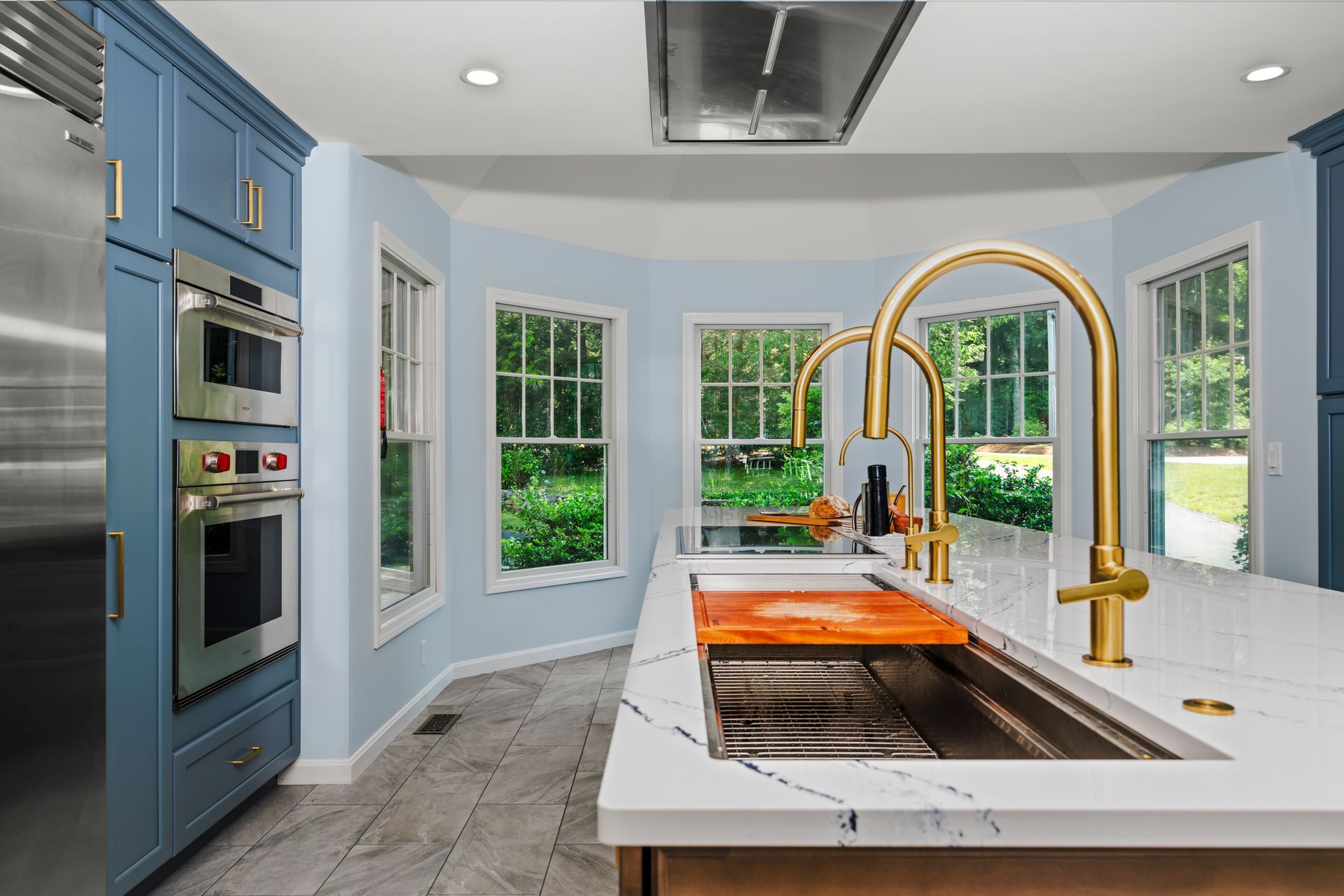 Bright kitchen with blue cabinets, white island, gold faucet, and large windows overlooking greenery