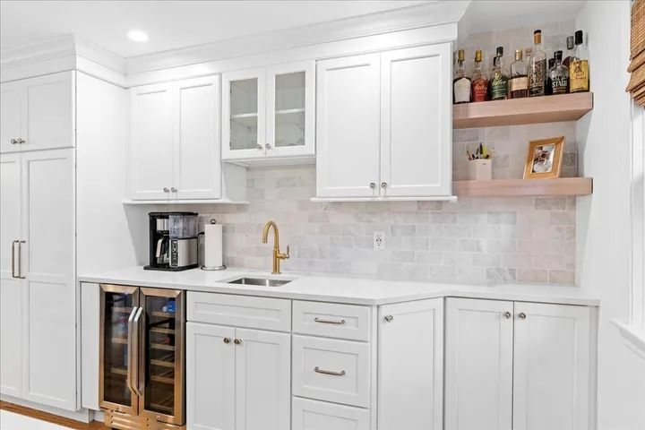 White kitchen with cabinets, sink, shelves with bottles, and wine cooler.