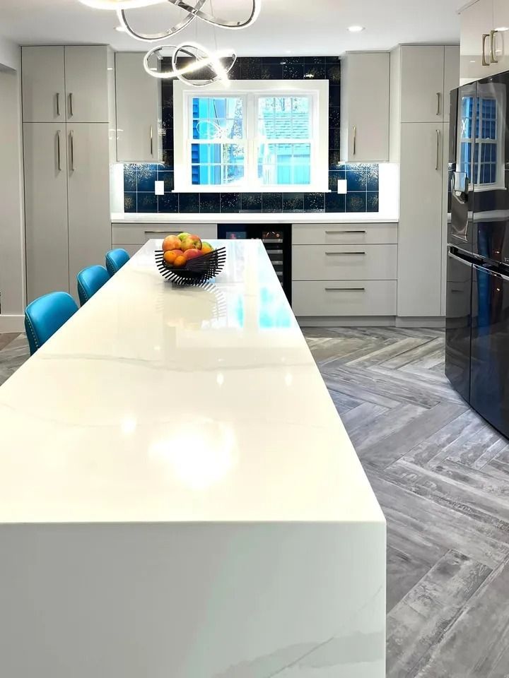 Modern kitchen with long white island, blue chairs, gray cabinets, and herringbone floor.