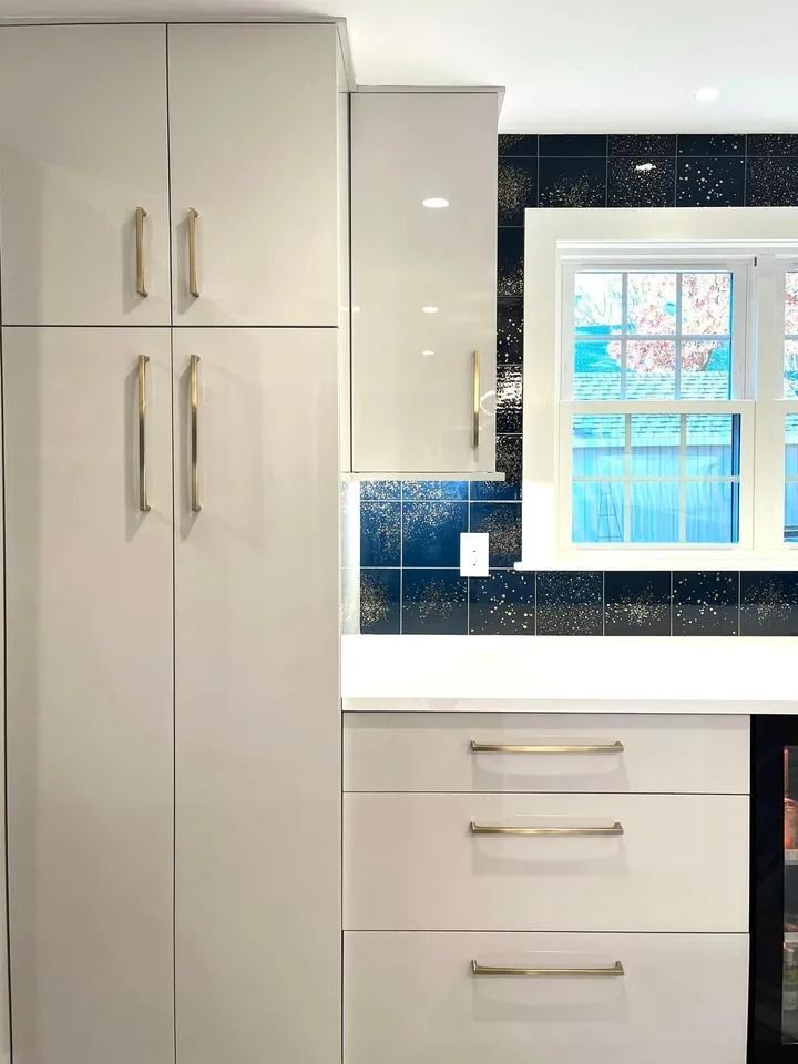 Modern kitchen with white cabinetry, gold handles, and dark blue backsplash near a window.