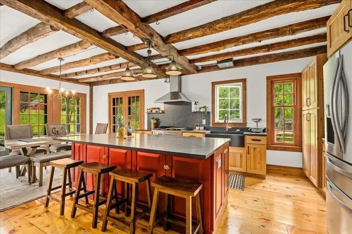 Rustic kitchen with red island, wood beams, and stools. Dining area and stainless steel appliances.