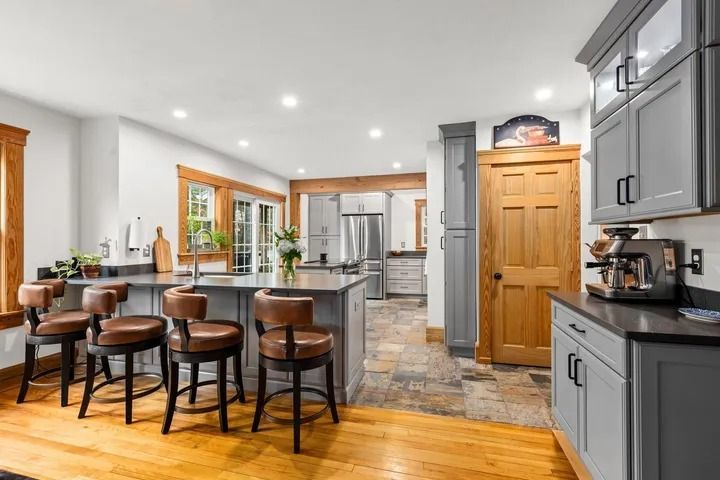 Open-concept kitchen with gray cabinets, wooden floors, and a breakfast bar with leather stools.