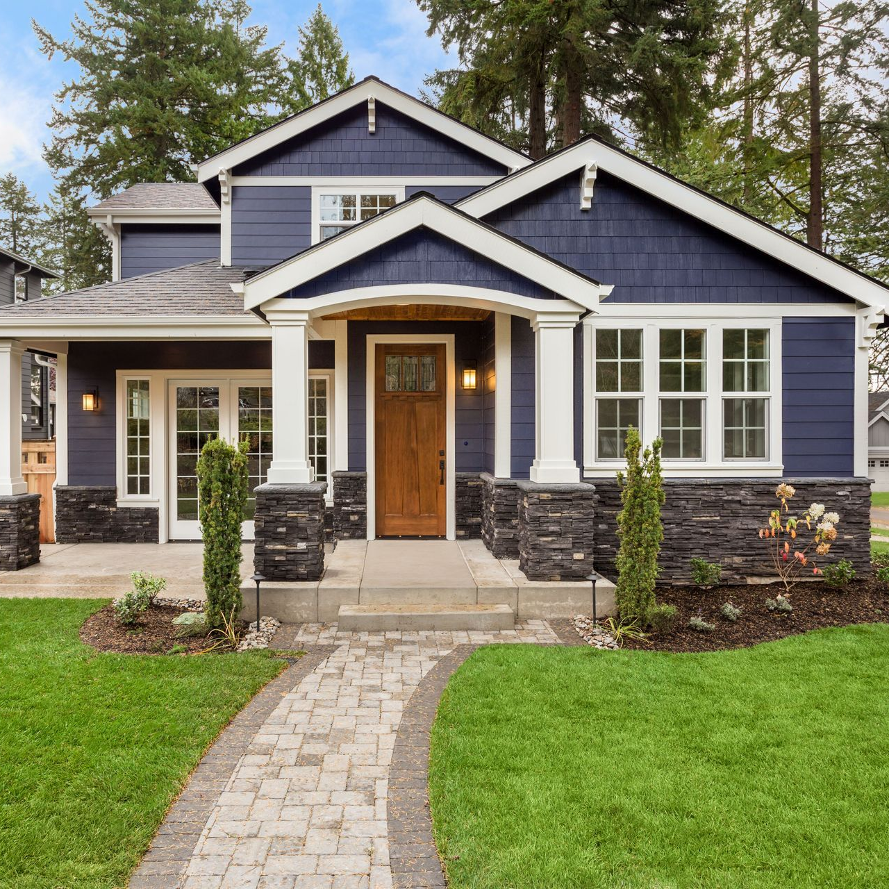 Blue house with white trim, brown door, stone accents, and paved path through a grassy yard.