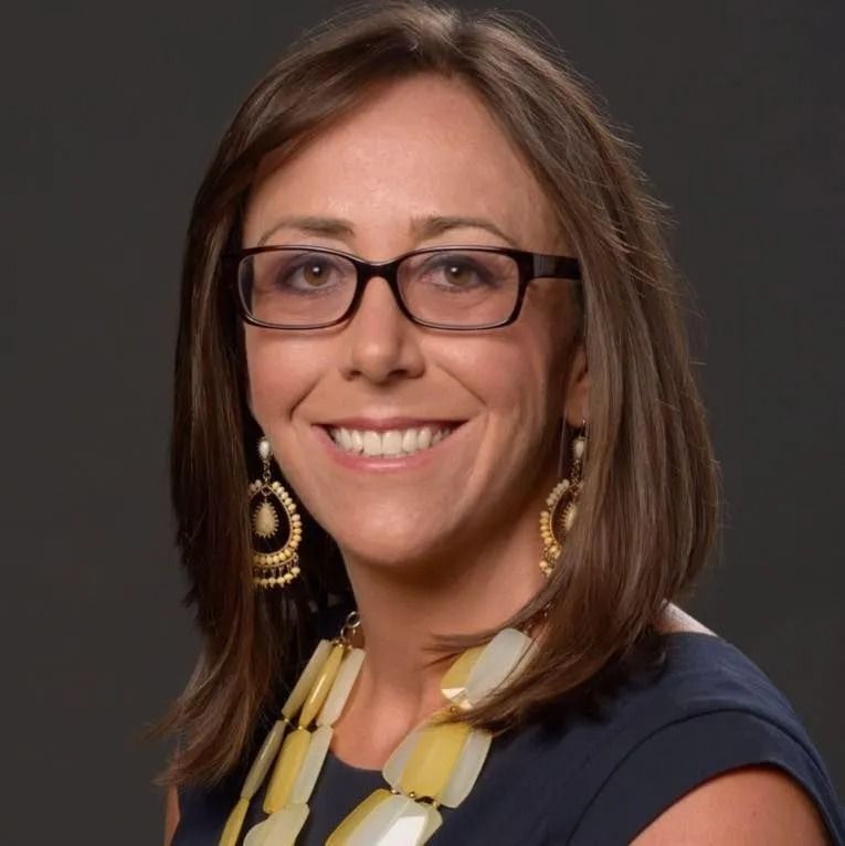 Woman with glasses, smiling, wearing a blue top and statement necklace.