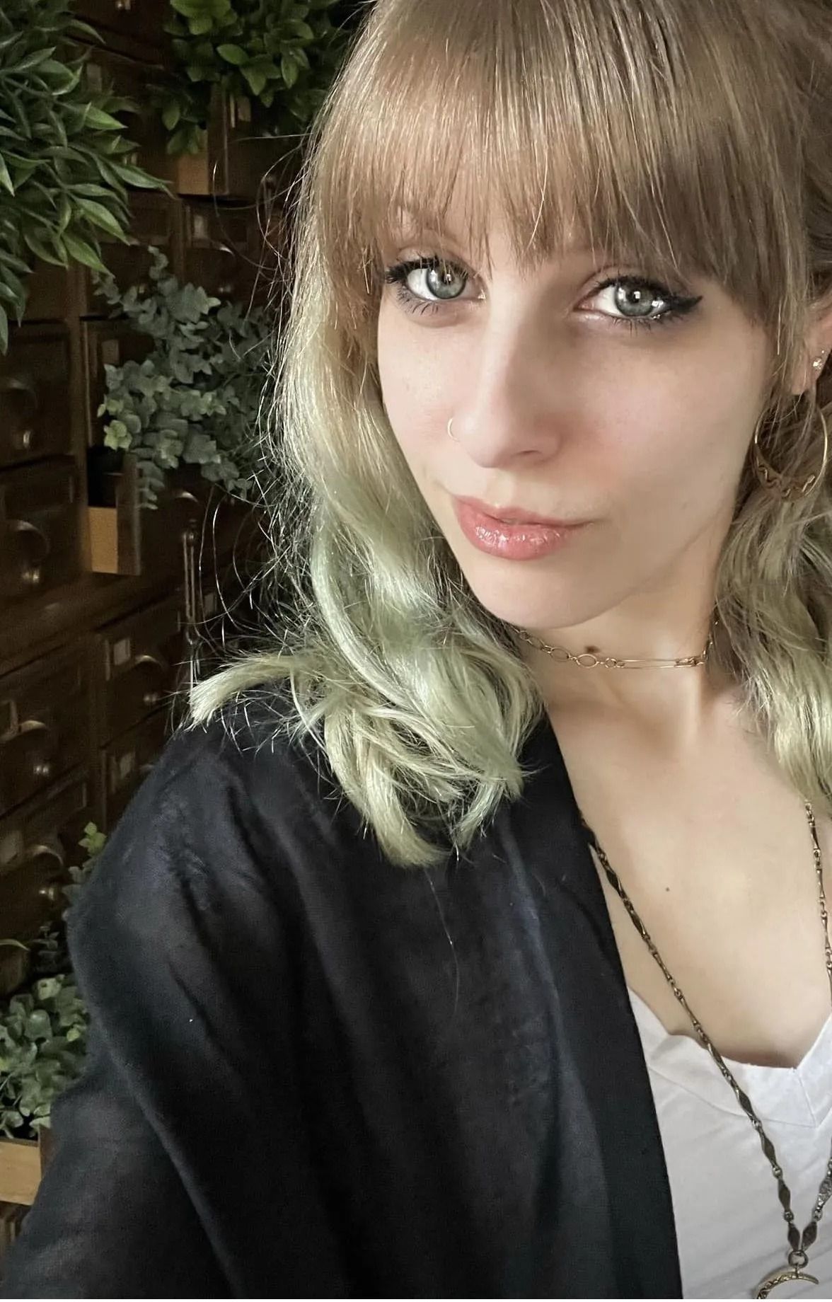Woman with green-tinted blonde hair, bangs, black cardigan, and white top poses against greenery.