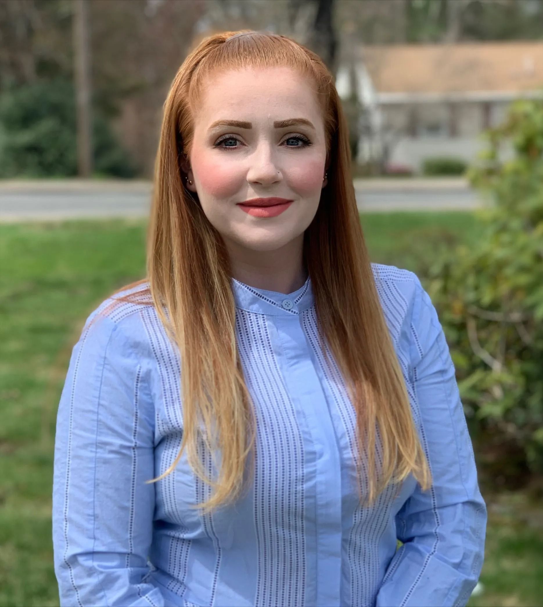 Woman with red hair in a blue striped shirt smiling outside in front of a green lawn and trees.