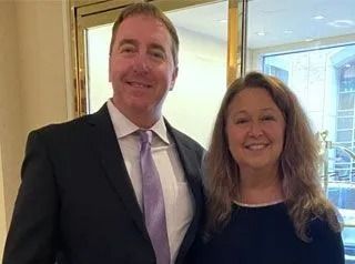 Man in suit and woman smiling, standing near a glass door.