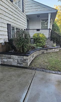 Front porch with black railing, stone planter wall, shrubs, and concrete walkway