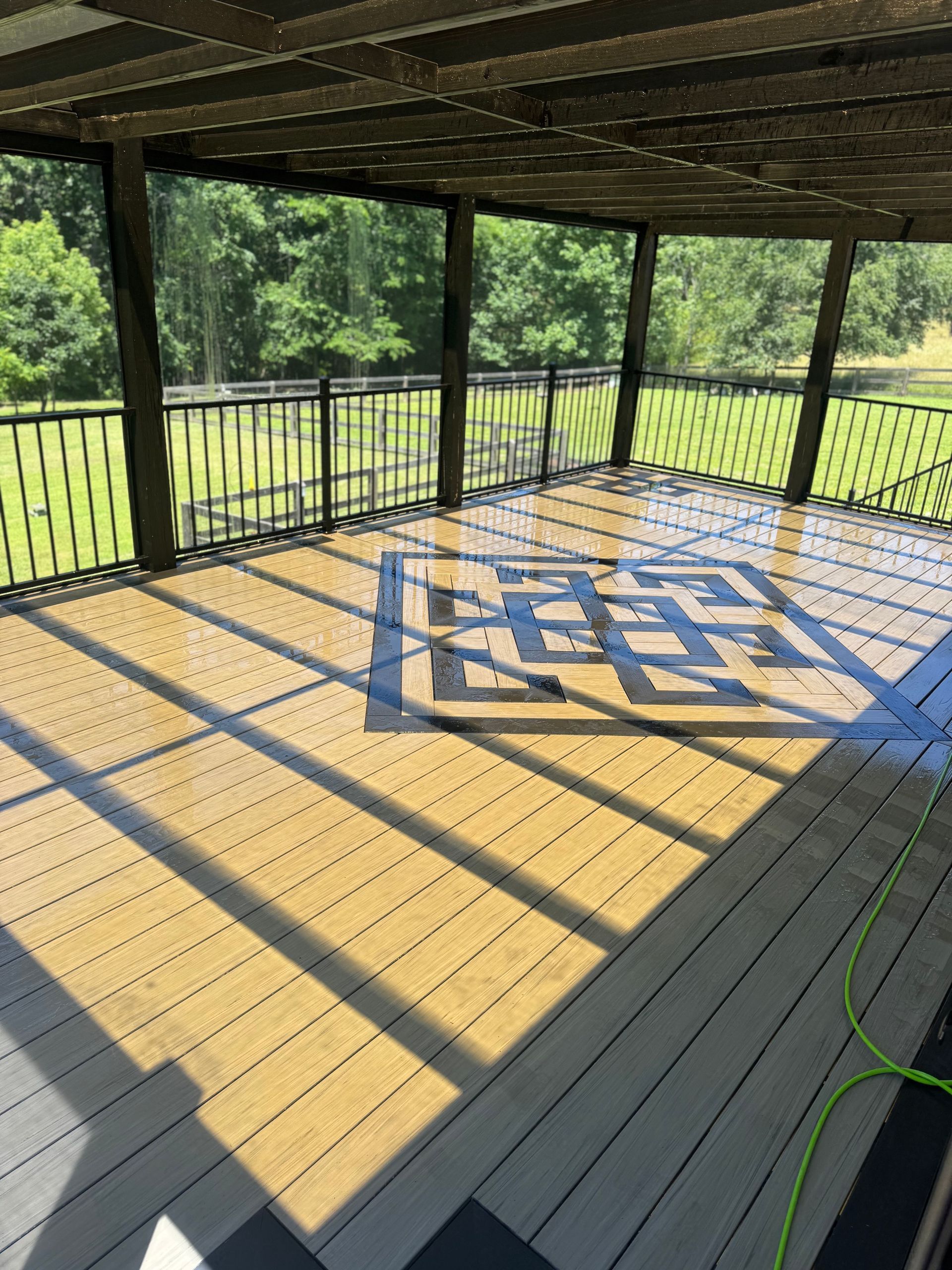 Wooden deck under a pergola with black railing, dappled sunlight, and a blue geometric rug or mat.