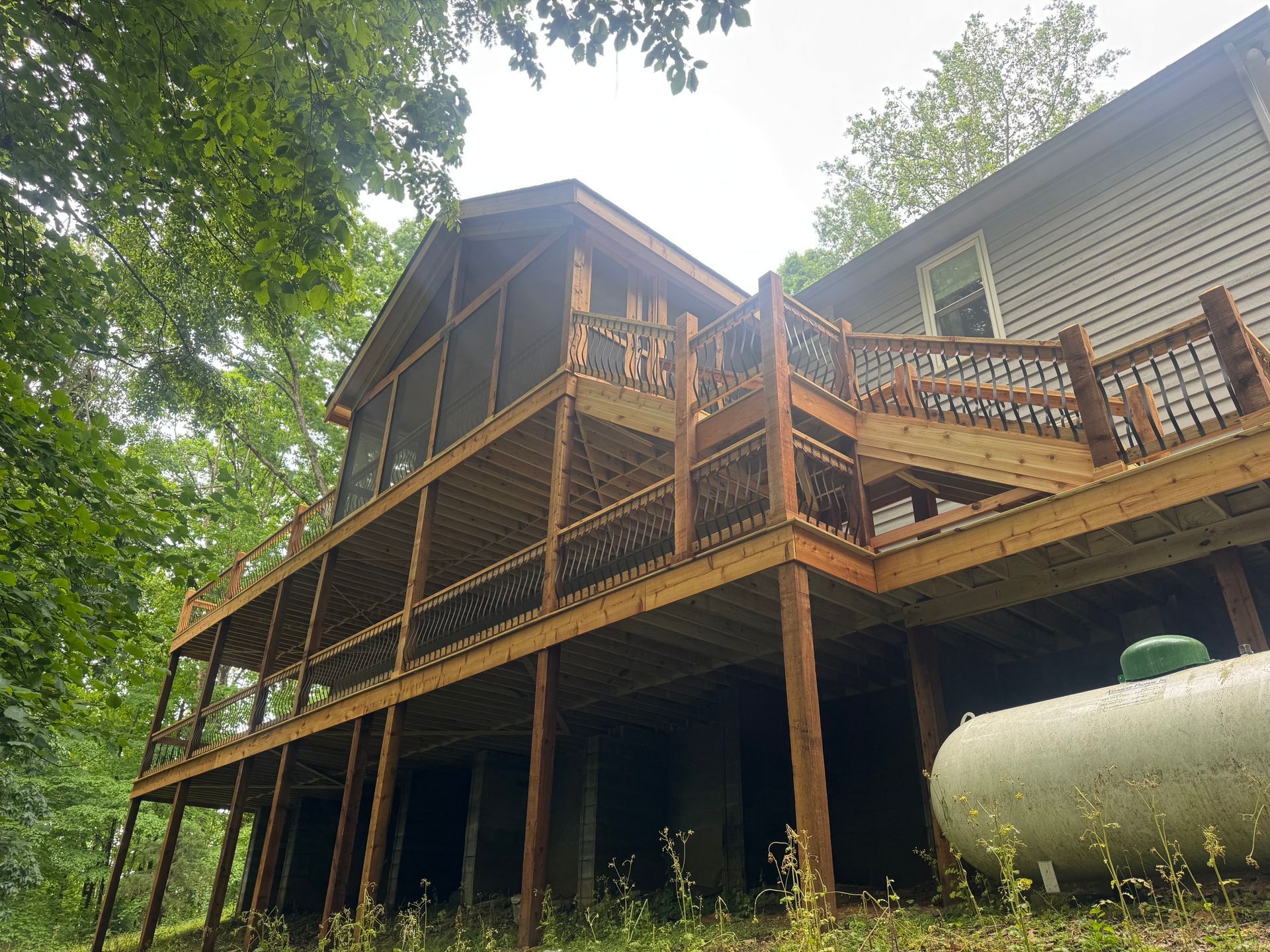 Wooden deck and screened porch on a raised house surrounded by trees, with a white propane tank below.