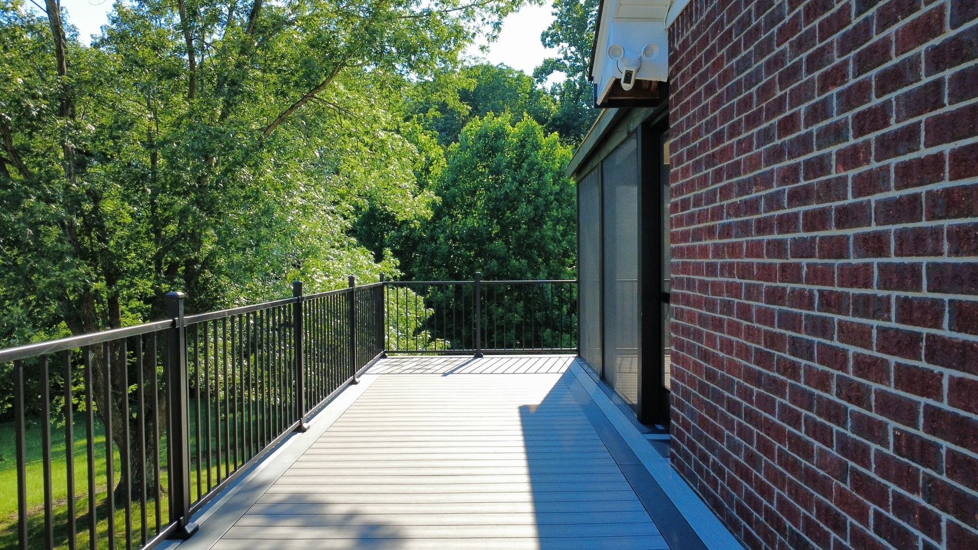 Brick building beside a sunlit wooden deck with black railing, overlooking green trees.