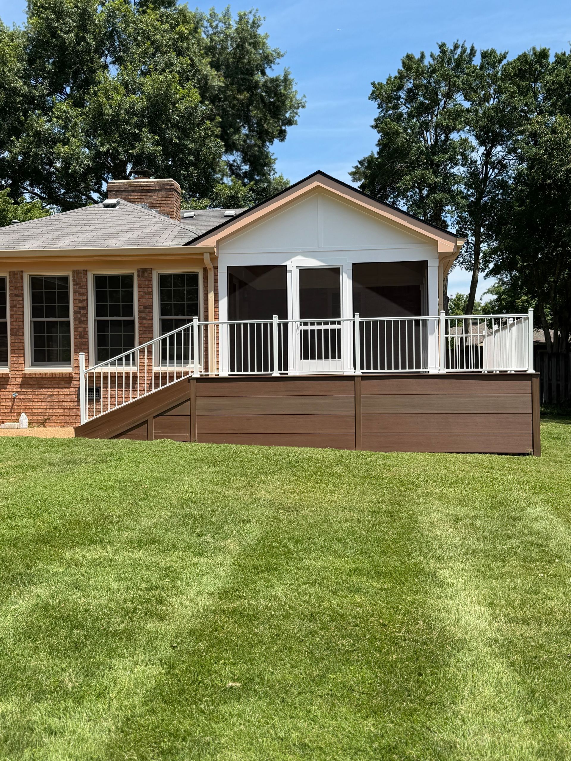 Single-story house with a white porch and brown deck overlooking a green lawn under trees.