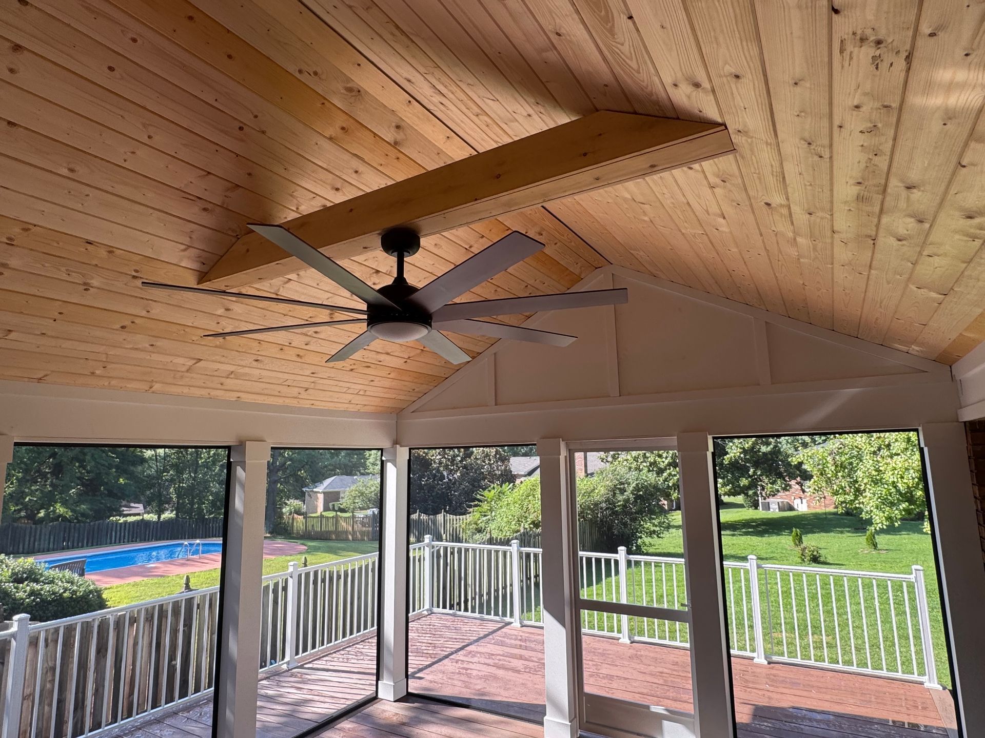 Covered patio with wood ceiling, ceiling fan, and large windows overlooking a backyard and pool.