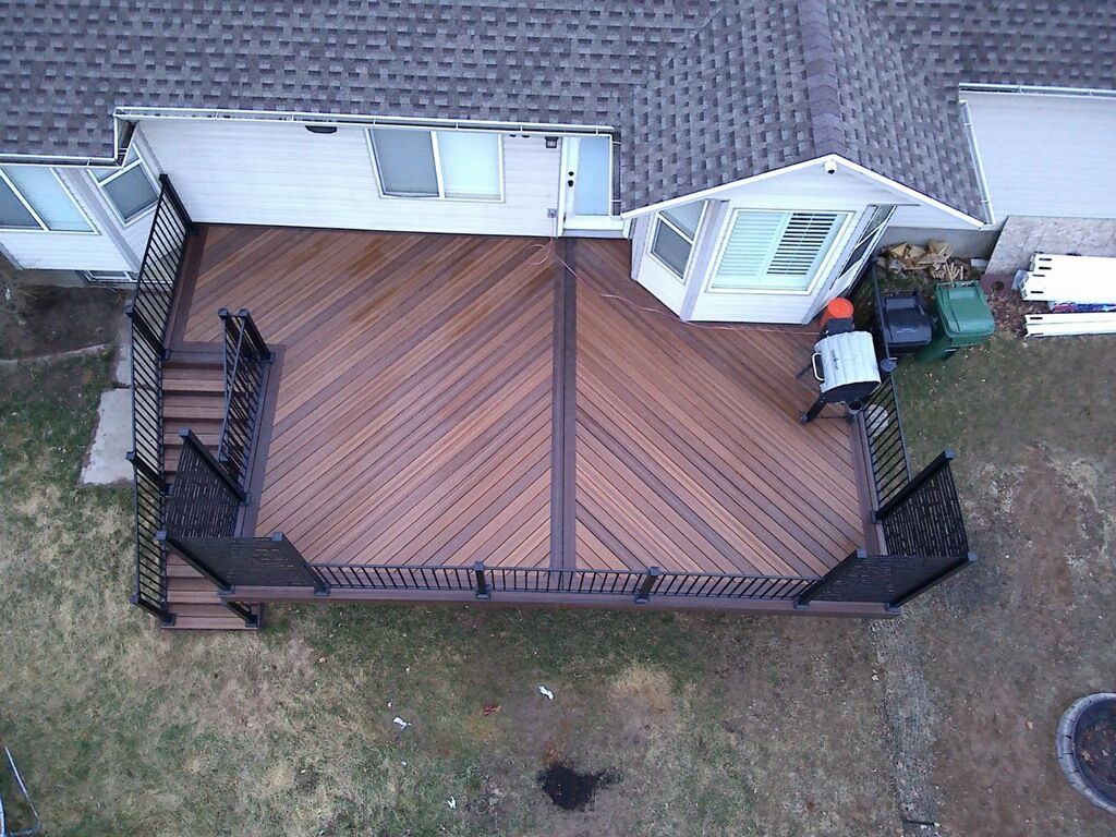 An aerial view of a brown wooden backyard deck with a diagonal pattern, black railings, stairs, and a grill.