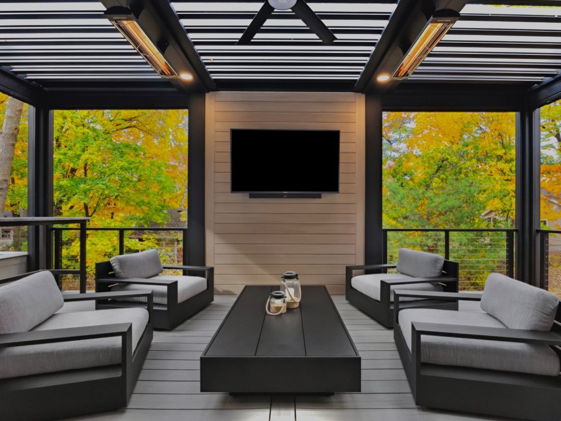 Modern outdoor patio with a flat-screen TV on a wood wall, central coffee table, and four gray chairs under a louvered roof.