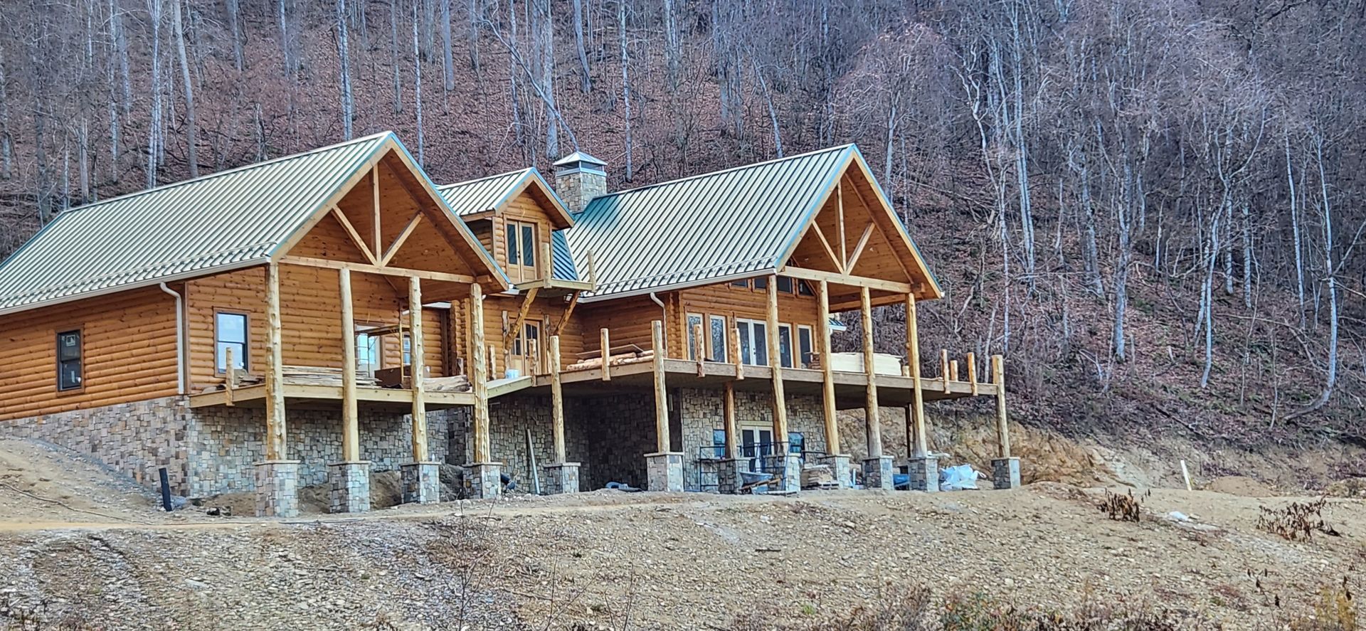 A partially constructed log cabin home with a stone foundation, set against a wooded hillside during the winter.
