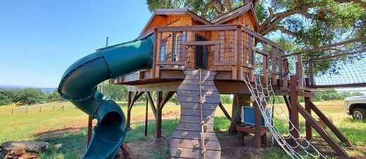 A backyard wooden treehouse with a green spiral slide, a climbing wall, and a rope ladder, set in a grassy field.
