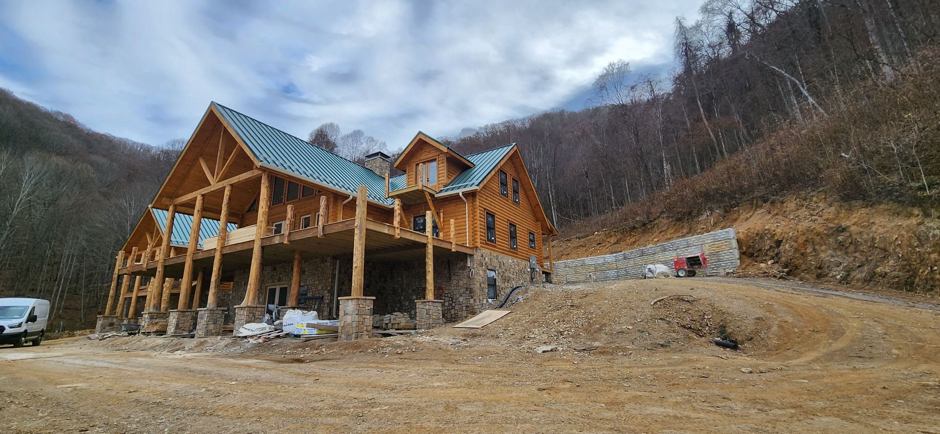 A large, unfinished wooden lodge with a stone foundation sits on a mountain hillside under a cloudy sky.