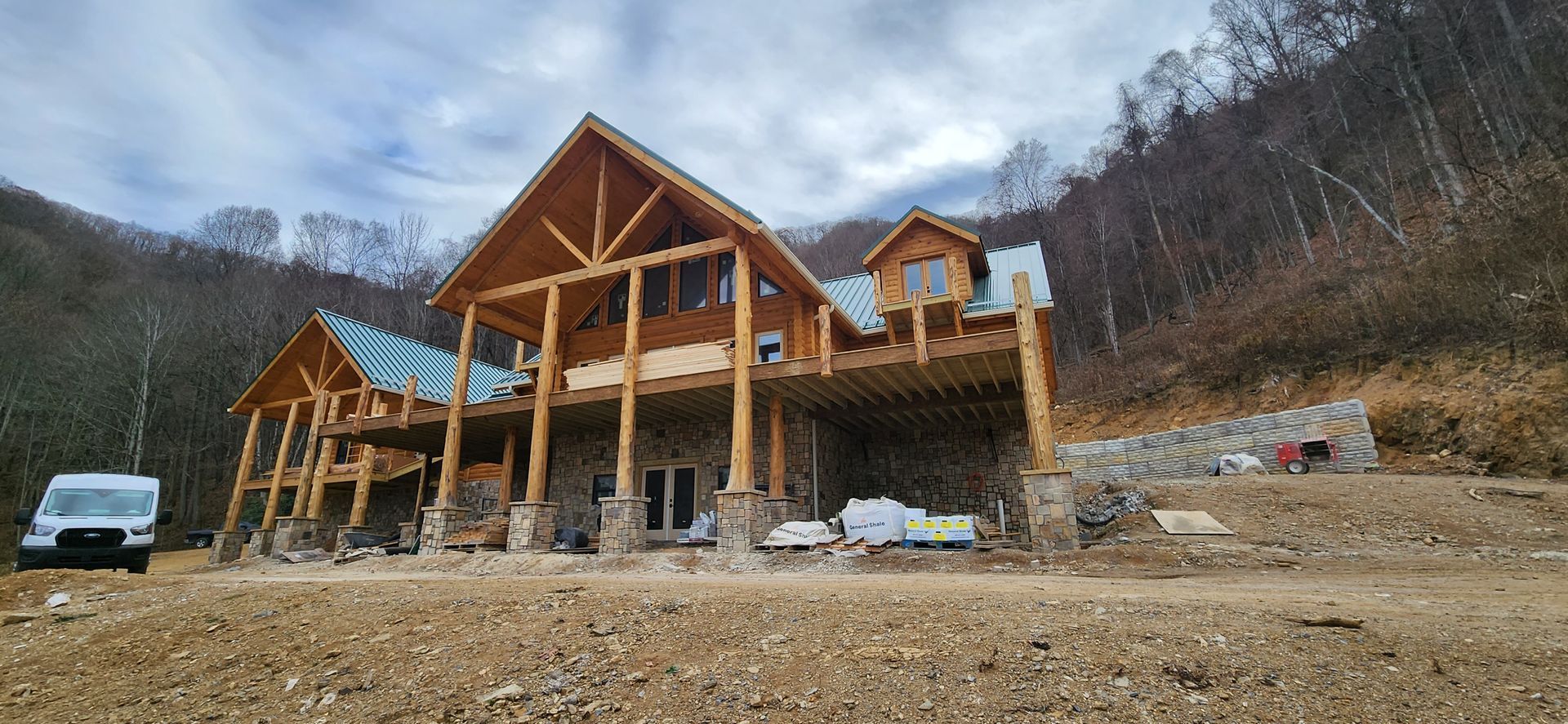 A large, under-construction log cabin with a stone base sits on a hillside near a white van under a cloudy sky.