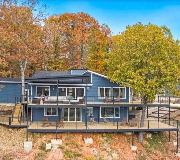 Blue lakeside house with wraparound deck, backed by autumn trees
