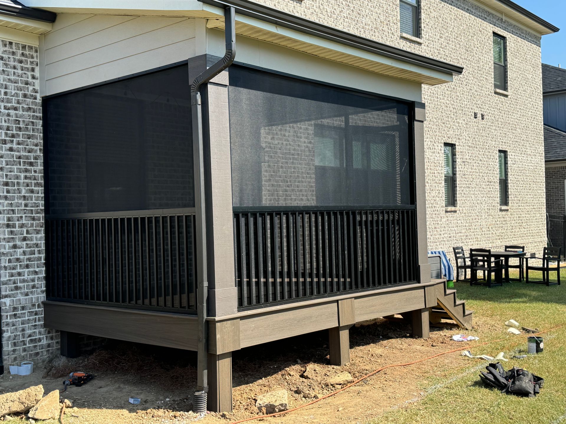A deck with black screened-in porch with roll-down blinds. Black railing and siding. Beige brick building behind.