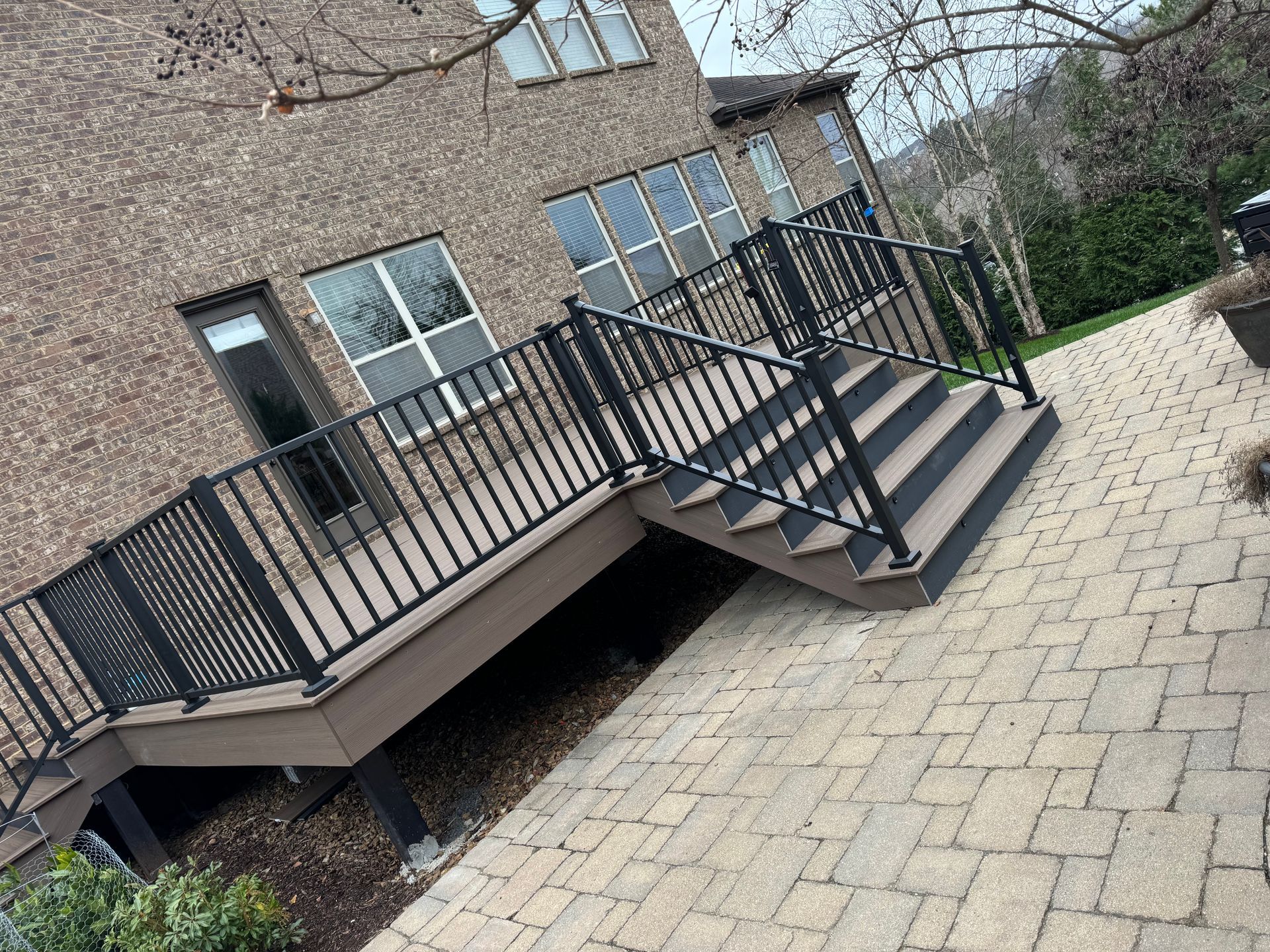 Deck with black railings and stairs, connected to a brick house, next to a paved pathway.