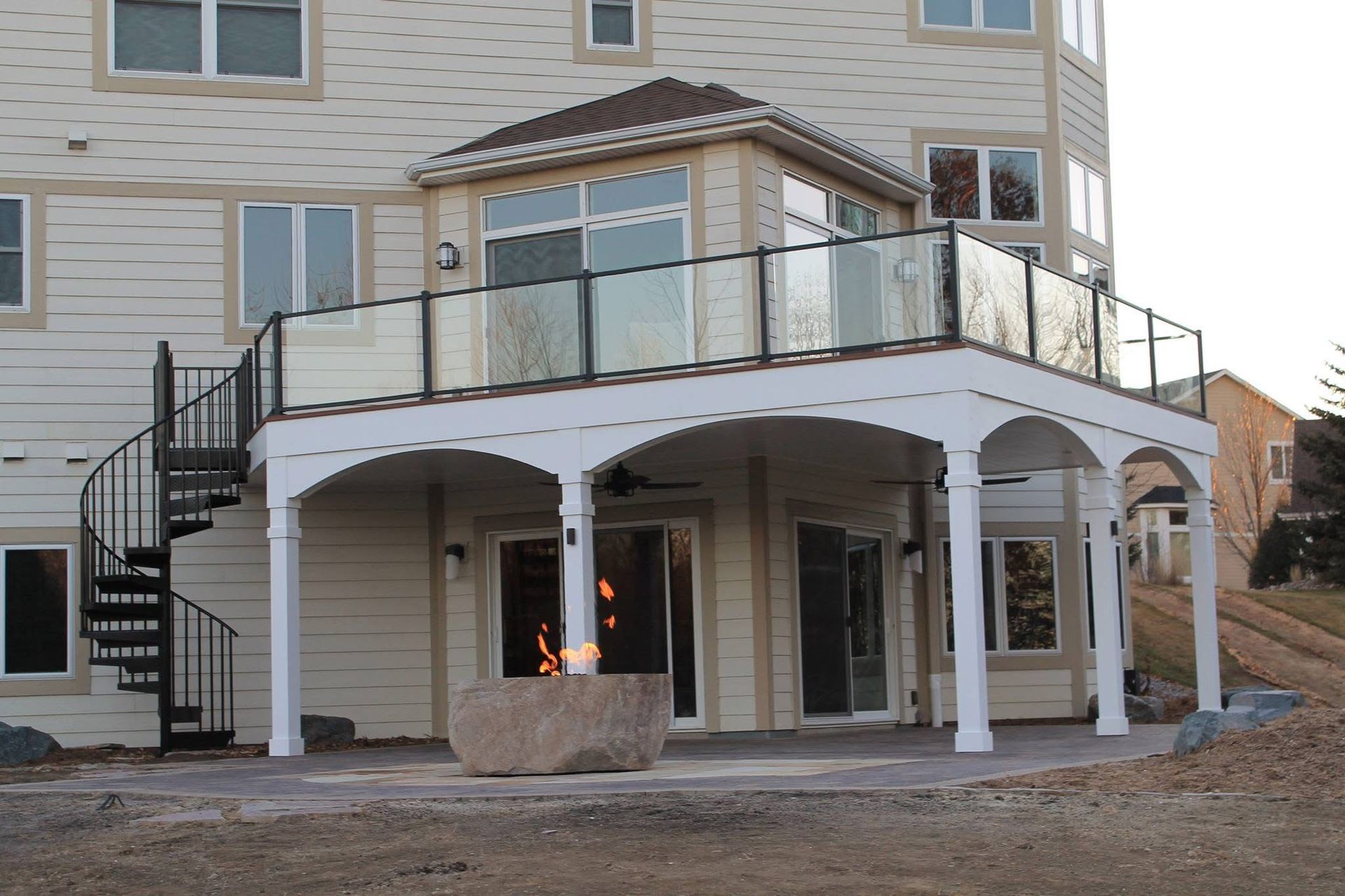 Two-story deck with glass railing, spiral staircase, and stone fire pit under an arched roof.