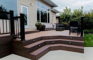Multi-level brown deck with black railing, steps, and built-in lighting, next to a house with large windows and a manicured lawn.